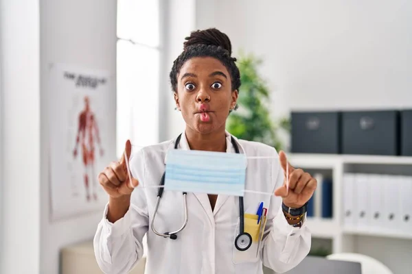 Young african american with braids wearing doctor uniform holding safety mask making fish face with mouth and squinting eyes, crazy and comical.