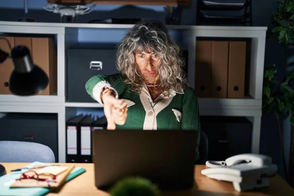 Middle age woman working at night using computer laptop looking unhappy and angry showing rejection and negative with thumbs down gesture. bad expression.