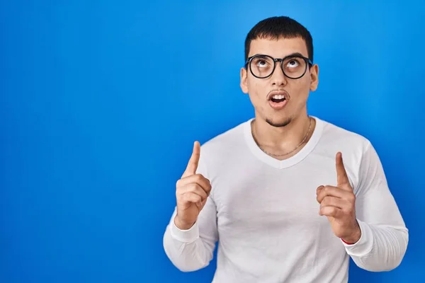 カジュアルな白いシャツと眼鏡を身に着けている若いアラブ人の男は驚きと驚きを見上げ 指と上げ腕で指して — ストック写真