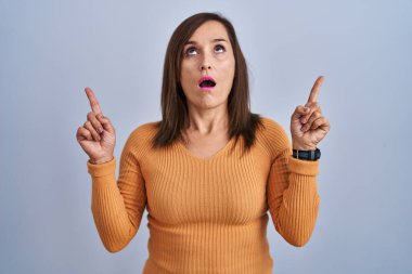 Middle age brunette woman standing wearing orange sweater amazed and surprised looking up and pointing with fingers and raised arms. 