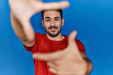 Young hispanic man with beard wearing red t shirt over blue background smiling making frame with hands and fingers with happy face. creativity and photography concept. 