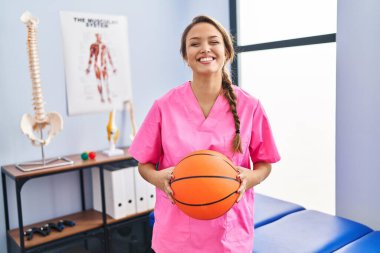 Young hispanic woman working at physiotherapy clinic holding basketball ball smiling with a happy and cool smile on face. showing teeth. 