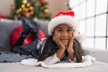 Adorable hispanic girl smiling confident lying on sofa by christmas tree at home