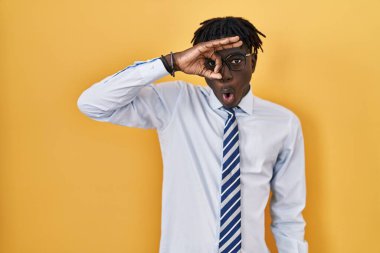 African man with dreadlocks standing over yellow background doing ok gesture shocked with surprised face, eye looking through fingers. unbelieving expression. 