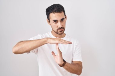 Handsome hispanic man standing over white background doing time out gesture with hands, frustrated and serious face 