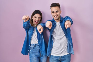 Young hispanic couple standing over pink background pointing to you and the camera with fingers, smiling positive and cheerful 