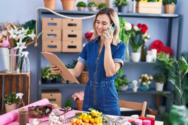 Young woman florist talking on smartphone reading clipboard at florist