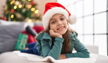 Adorable hispanic girl smiling confident lying on sofa by christmas tree at home