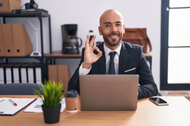 Middle age bald man working at the office doing ok sign with fingers, smiling friendly gesturing excellent symbol 