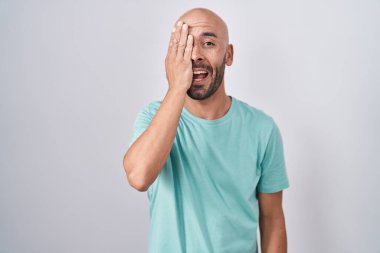 Middle age bald man standing over white background covering one eye with hand, confident smile on face and surprise emotion. 