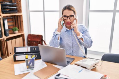 Young hispanic business woman working at the office talking on two phones clueless and confused expression. doubt concept. 