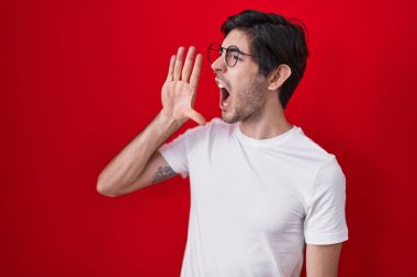 Young hispanic man standing over red background shouting and screaming loud to side with hand on mouth. communication concept. 