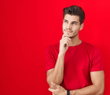 Elleri çenesinde kırmızı arka planda dikilmiş soru soran, düşünceli bir İspanyol genç. Düşünceli bir yüzle gülümsüyor. şüphe kavramı. 