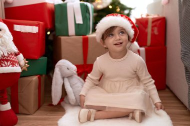 Sevimli kız, kendine güvenerek gülümsüyor Noel ağacının ve hediyelerin yanında oturuyor.