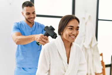 Latin erkek ve kadın fizyoterapi üniforması giyiyorlar boyuna masaj yapıyorlar güzellik merkezinde perküsyon tabancası kullanıyorlar.