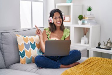 Hispanic young woman using laptop at home smiling and looking at the camera pointing with two hands and fingers to the side. 