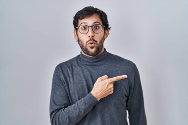 Handsome latin man standing over isolated background surprised pointing with finger to the side, open mouth amazed expression. 