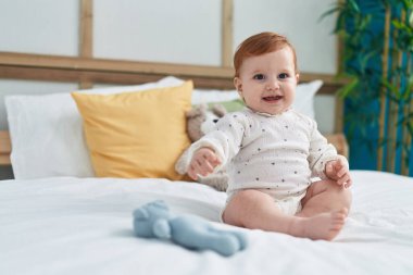 Sevimli kızıl saçlı bebek gülümsüyor. Yatak odasında kendinden emin bir şekilde oturuyor.