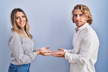 Young couple standing over blue background pointing aside with hands open palms showing copy space, presenting advertisement smiling excited happy 
