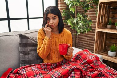 Young hispanic woman sitting on the sofa drinking a coffee at home pointing to the eye watching you gesture, suspicious expression 