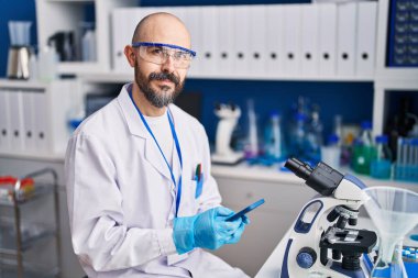 Genç kel bilim adamı laboratuvarda akıllı telefon kullanarak gülümsüyor.