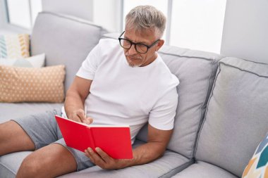 Orta yaşlı, kır saçlı, kitap okuyan adam evdeki kanepede oturuyor.