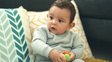 Adorable hispanic baby holding toy sitting on sofa at home