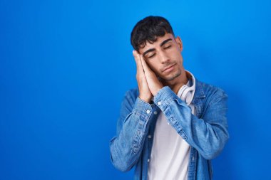 Genç İspanyol adam mavi arka planda ayakta uyur. Yorgun düşler kurar, ellerle poz verir ve gözleri kapalı gülümserdi.. 