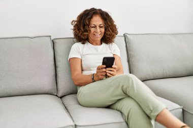 Orta yaşlı İspanyol kadın evde akıllı telefon kullanarak gülümsüyor.