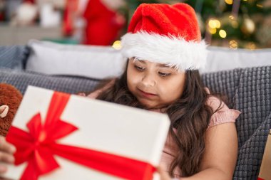 Plus size hispanic girl unpacking gift sitting on sofa by christmas tree at home
