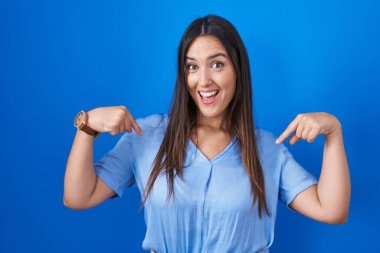 Genç esmer kadın mavi arka planda duruyor. Kendine güveni tam görünüyor. Kendini gururlu ve mutlu bir şekilde gösteriyor.. 