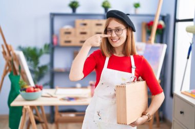 Young redhead woman at art studio holding art case pointing with hand finger to face and nose, smiling cheerful. beauty concept 