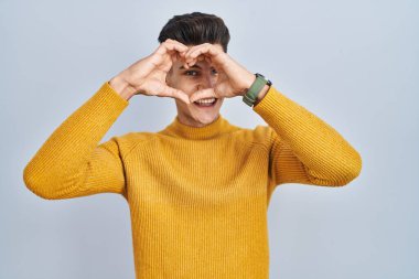 Mavi arka planda duran, elleri ve parmaklarıyla kalp şeklinde gülümseyen İspanyol genç adam tabelanın içinden bakıyor. 