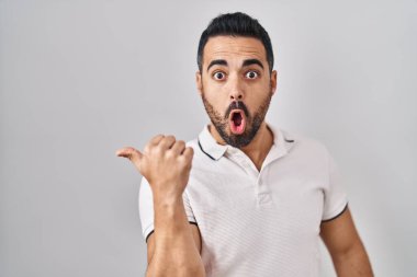 Beyaz arka planda sıradan kıyafetler giyen, sakallı, genç İspanyol bir adam elini yana uzatmış, ağzı açık, şaşkın bir ifade sergiliyor.. 