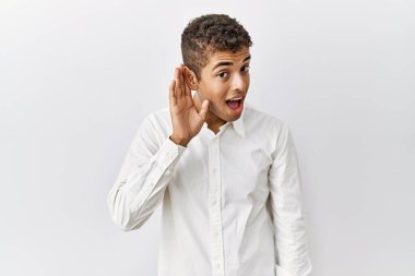 Young handsome hispanic man standing over isolated background smiling with hand over ear listening an hearing to rumor or gossip. deafness concept. 
