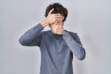 Young man standing over isolated background covering eyes and mouth with hands, surprised and shocked. hiding emotion 