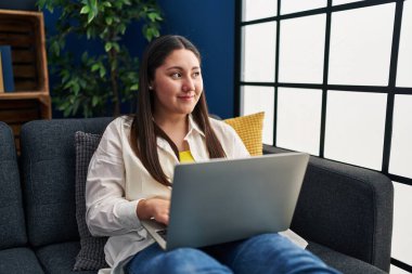 Genç İspanyol kadın evdeki koltukta dizüstü bilgisayar kullanıyor.