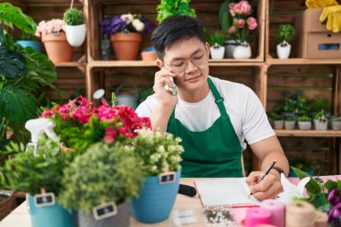 Young chinese man florist talking on smartphone writing on notebook at flower shop