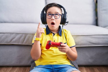 Young hispanic kid playing video game holding controller wearing headphones amazed and surprised looking up and pointing with fingers and raised arms. 