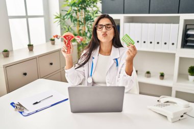 Young hispanic doctor woman holding anatomical female genital organ and birth control pills looking at the camera blowing a kiss being lovely and sexy. love expression. 