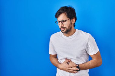 Handsome latin man standing over blue background with hand on stomach because nausea, painful disease feeling unwell. ache concept. 