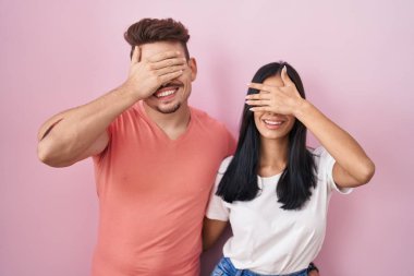 Young hispanic couple standing over pink background smiling and laughing with hand on face covering eyes for surprise. blind concept. 
