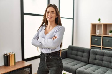 Genç İspanyol kadın gülümsüyor. Kollarını kavuşturup evde duruyor.