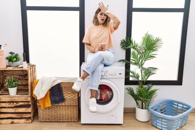 Young hispanic woman drinking coffee waiting for washing machine at laundry room surprised with hand on head for mistake, remember error. forgot, bad memory concept. 