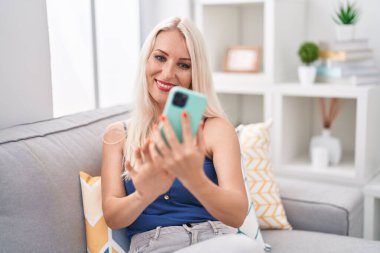 Evdeki kanepede oturan akıllı telefon kullanan genç sarışın kadın.