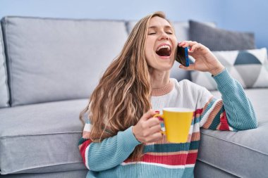 Young blonde woman talking on smartphone drinking coffee at home