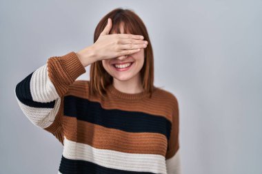 Young beautiful woman wearing striped sweater over isolated background smiling and laughing with hand on face covering eyes for surprise. blind concept. 