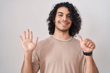 Beyaz arka planda duran kıvırcık saçlı İspanyol adam altı numaralı parmaklarını gösterip işaret ederken kendinden emin ve mutlu gülümsüyor.. 