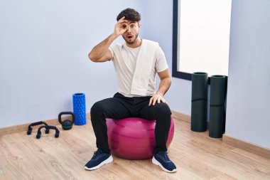 Sakallı İspanyol bir adam yoga odasında pilatese oturmuş şaşkın şaşkın hareketler yapıyor. Gözleri parmaklarından bakıyor. İnançsız ifade. 