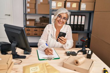 Orta yaşlı kadın ecommerce iş işçisi. Ofisteki panoya akıllı telefon yazısıyla yazıyor.